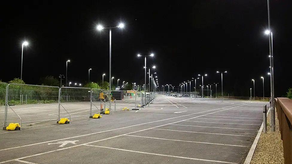 A large car park with multiple street lights, shown at night with the lights on and a barrier down the middle