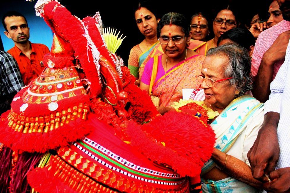 An older woman holding a brightly coloured red display, surrounded by other women