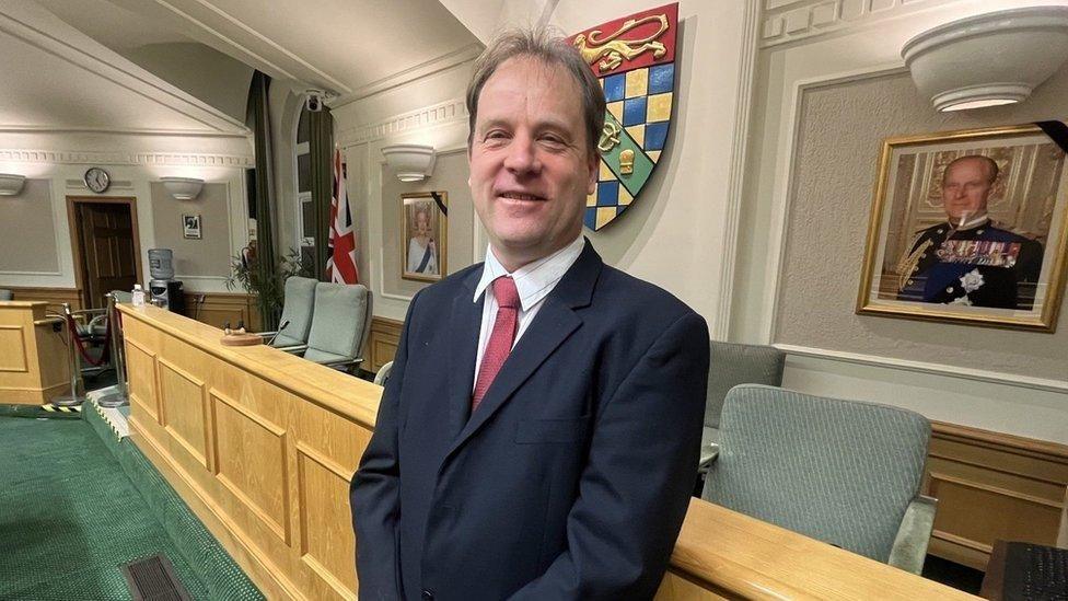 South Kesteven Council leader Ashley Baxter wearing a dark suit and red tie  stood in the council chamber in front of the council's coat of arms and the union flag and portraits of the late Queen and Prince Philip