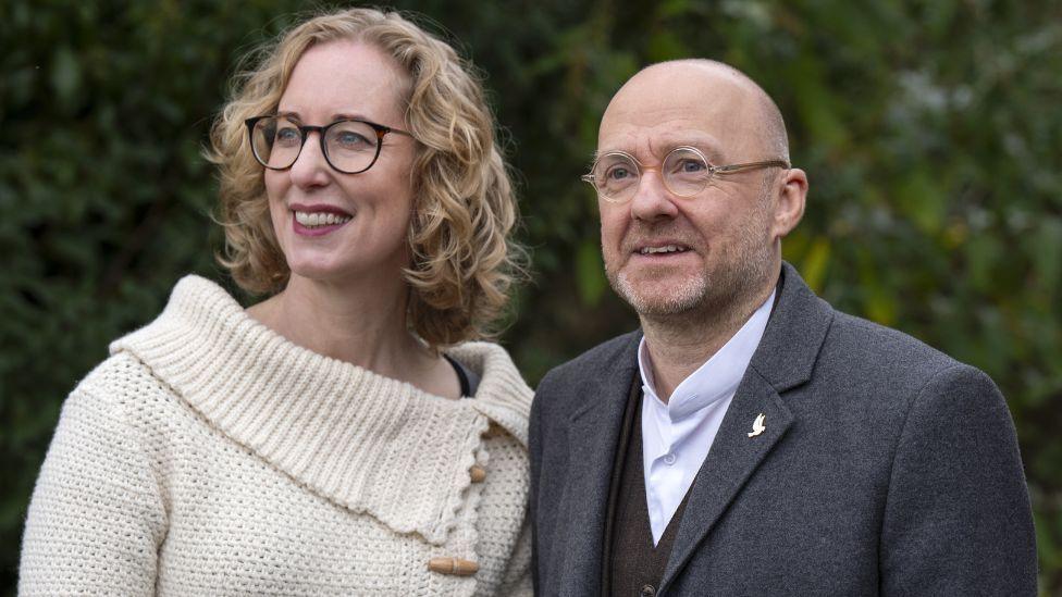 Slater, a woman with blonde curly hair and black-rimmed glasses, stands next to Harvie, a bald man with glasses. Slater is wearing a cream-coloured knitted jumper. Harvie is wearing a charcoal suit and lilac shirt. There are trees in the background.