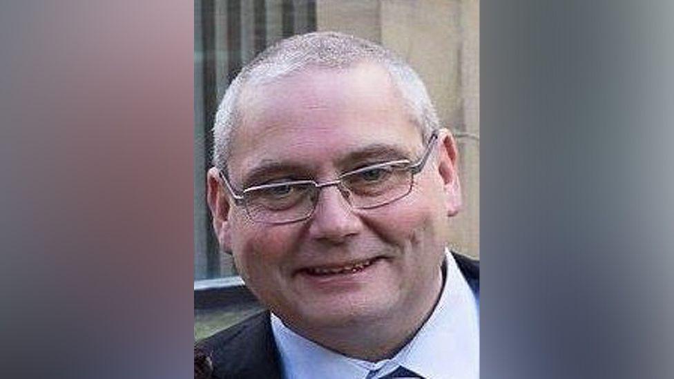 David Daintree smiles at the camera in a suit and shirt and tie. He has short grey hair and silver rimmed glasses. 