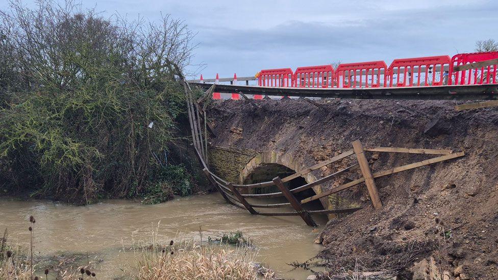 A damaged culvert under the A1 southbound over Billing Brook, Water Newton. It shows a damaged traffic barrier tipping over a muddy bank leading to the brick-built arched culvert opening. A wooden fence has tumbled down the bank and is tipped into the water, which is muddy with silt. There are shrubs to the left of the brook. A red crash barrier and red and white cones run along the edge of the road above the culvert.