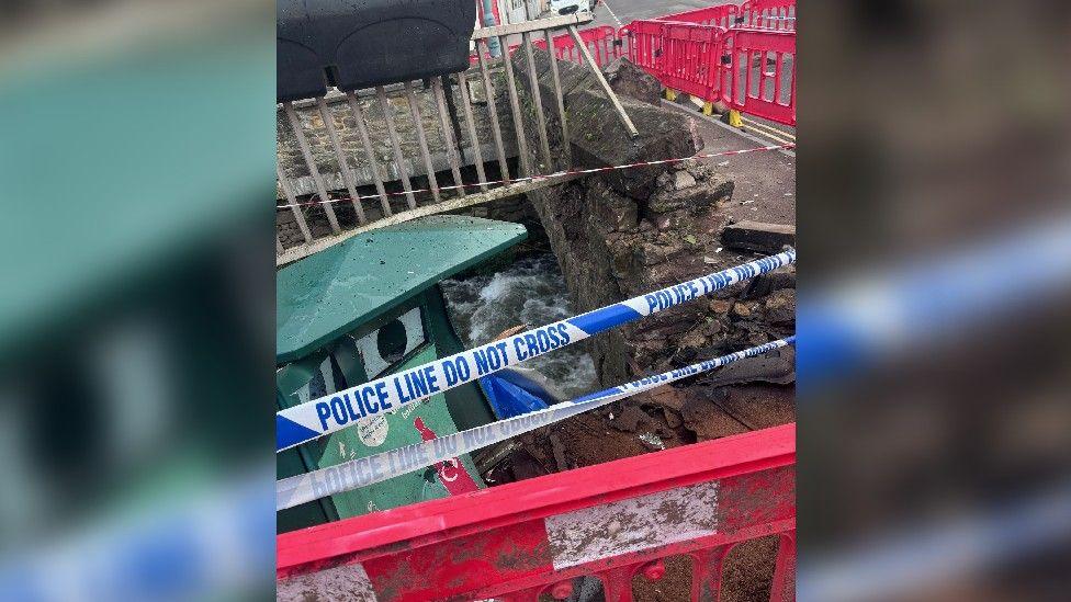 A partially destroyed stone bridge that has been hit by a car, with damaged metal railings, bright orange barricades, and a police line over the water below