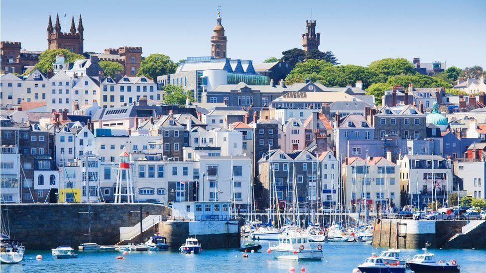 A photo which shows Guernsey. There is blue water in the forefront with boats dotted on the harbour. Behind it are different coloured and shaped buildings.