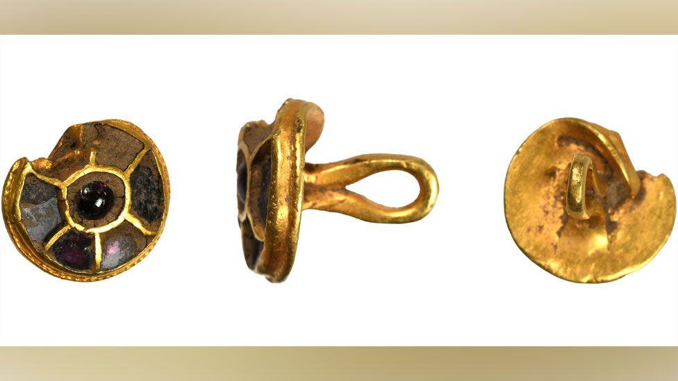 Three views of an Anglo-Saxon gold and red stone button or stud, slightly damaged on its top edge. The first view on the left shows the front, which is round and has six compartments on its face, which were inlaid with red stones - some still existing. The middle view shows its side and the gold loop attached to its back. The third view on the right is of its back, which is entirely gold and has the loop facing to the viewer. 