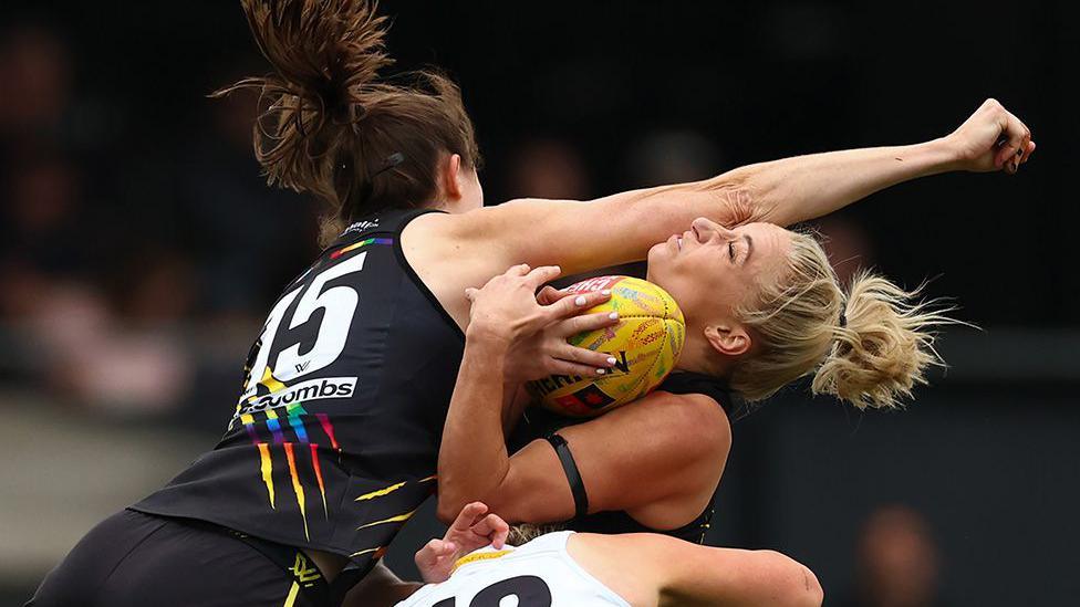 Two Australian rules football players contest possession, one holding the ball tightly while being tackled by an opponent in a black jersey during a physical play.