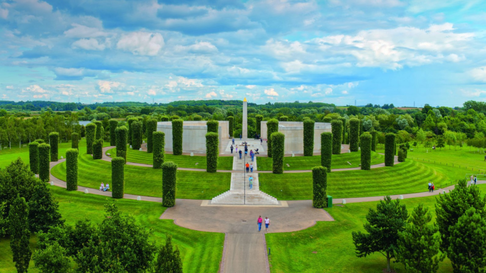 A memorial in the middle of wooded countryside. A white monument can be seen in the distance with white walls on either side and white steps leading up to it. Footpaths encircle the memorial along with a number of vertical hedges.