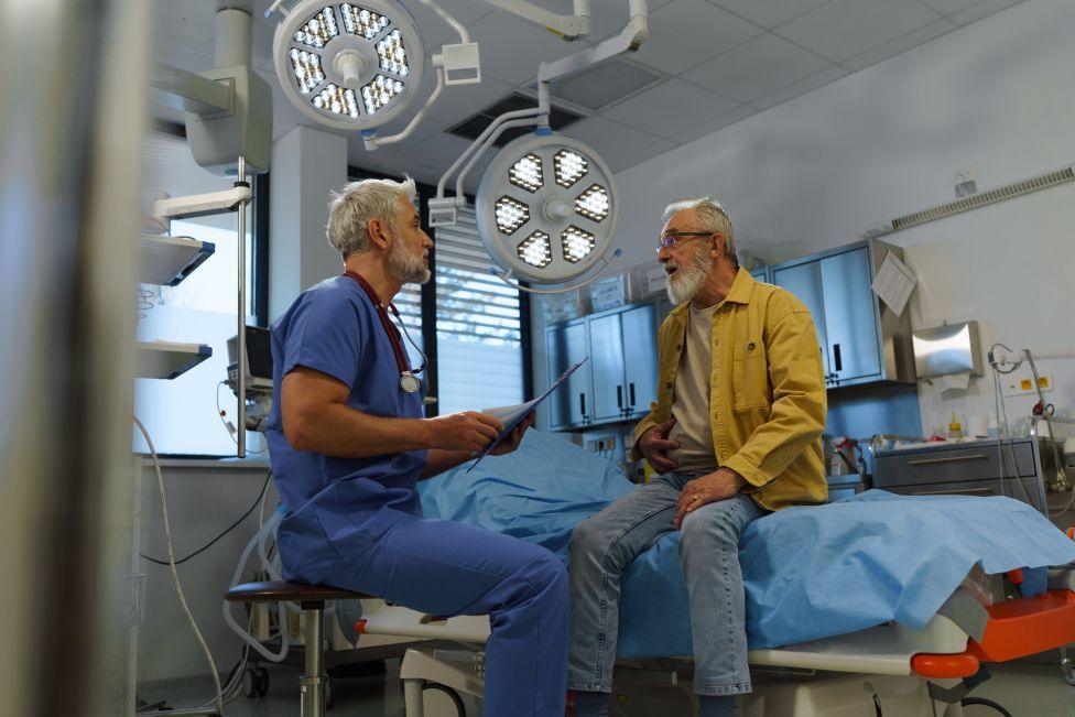 Doctor dressed in blue scrubs talks to older patient dressed in jeans and yellow jacket sat on a hospital bed.