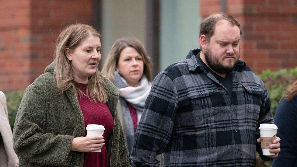 Sarah and Gary Andrews holding white takeaway coffee cups as they walk past a red brick building. There are other people around them walking in the same direction. Sarah, with long blonde hair, wears a dark red dress and olive green coat. Gary, who has a beard, wears a dark t-shirt and grey checked shirt