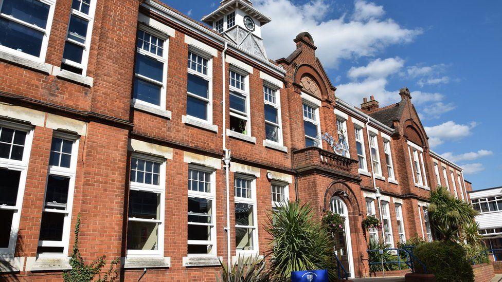 A wide, two-storey school building at Plume Academy, made out of red brick and featuring lots of windows with white panes. There are various plants in front of the building and the picture has been taken on a sunny day.