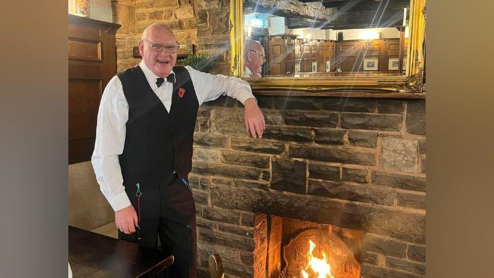 Dennis Duckhouse, a bald man, stands leaning against a brick fireplace. He is wearing a black waistcoat and whit shirt, along with a black bow tie. On the wall behind him is a gold mirror. He is smiling