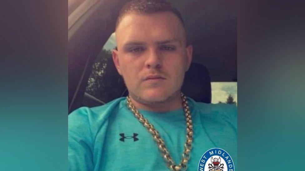 A man with short hair and wearing a gold chain and a green T-shirt is sitting in a car.