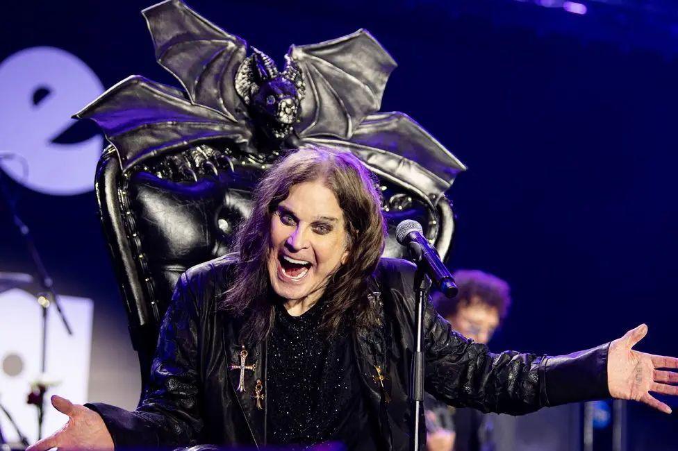 A seated Ozzy Osbourne wears dark eye make-up and a dark leather jacket at the Villa Park gig. His throne has a bat at the head.