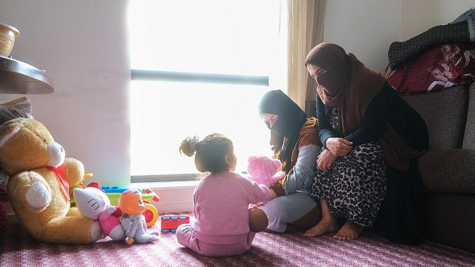 Two women with faces covered and a small child sit on a carpeted floor in a softly lit room with a large window. One woman holds a pink stuffed animal, another sits nearby, and a a small child in pink pyjamas faces away from the camera, playing with toys. Various stuffed animals and toys are scattered around them, creating a cozy and playful atmosphere.

