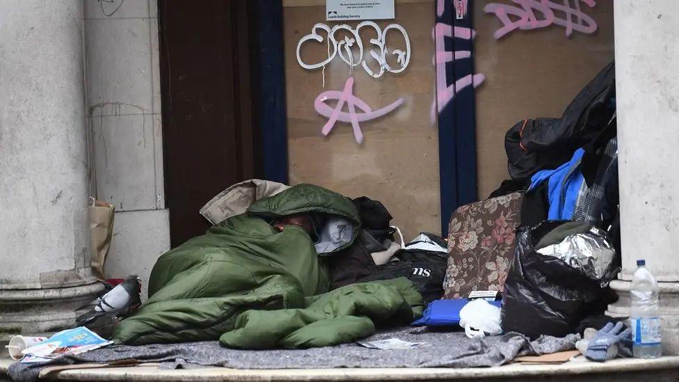 A person inside a green sleeping bag in a disused shop doorway