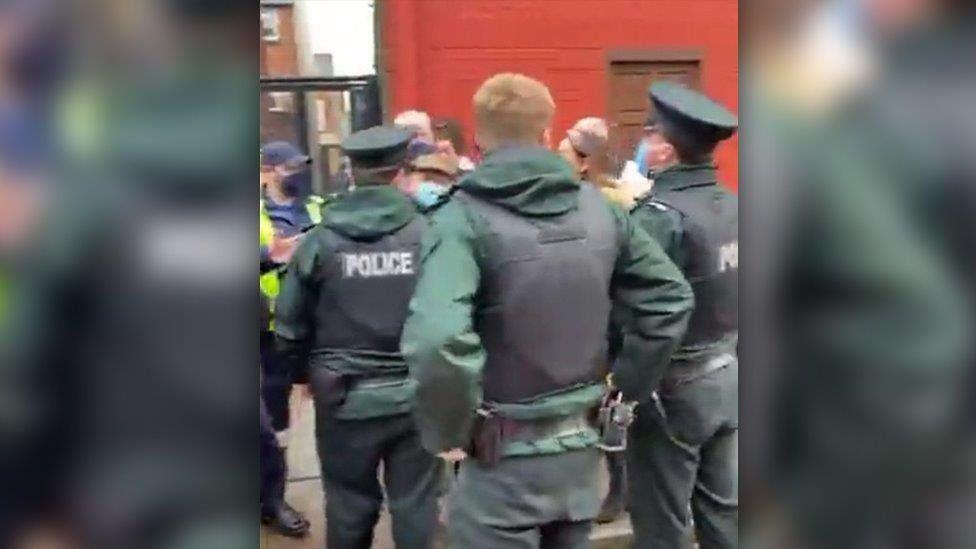 A group of police officers and people are standing in a street. The officers are standing away from the camera. People are wearing face masks.