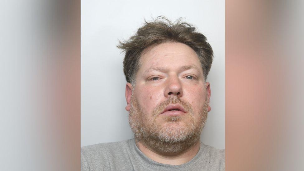 A police mugshot of a man aged around 40, with brown untidy hair and a short beard. He is wearing a grey t-shirt and staring into the camera directly.