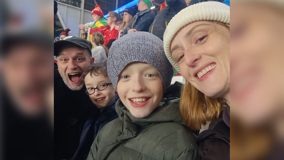 Selfie picture of John Foster (far left) with his two children (centre) and his wife (far right) at the football game all smiling into the lens. 