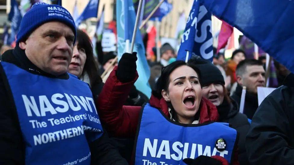 A picture of an NASUWT picket line, in the forefront are a man and woman wearing blue NASUWT bibs, with the woman waving a NASUWT flag and the man wearing a union hat. They are surrounded by other union members, many of them waving flags 