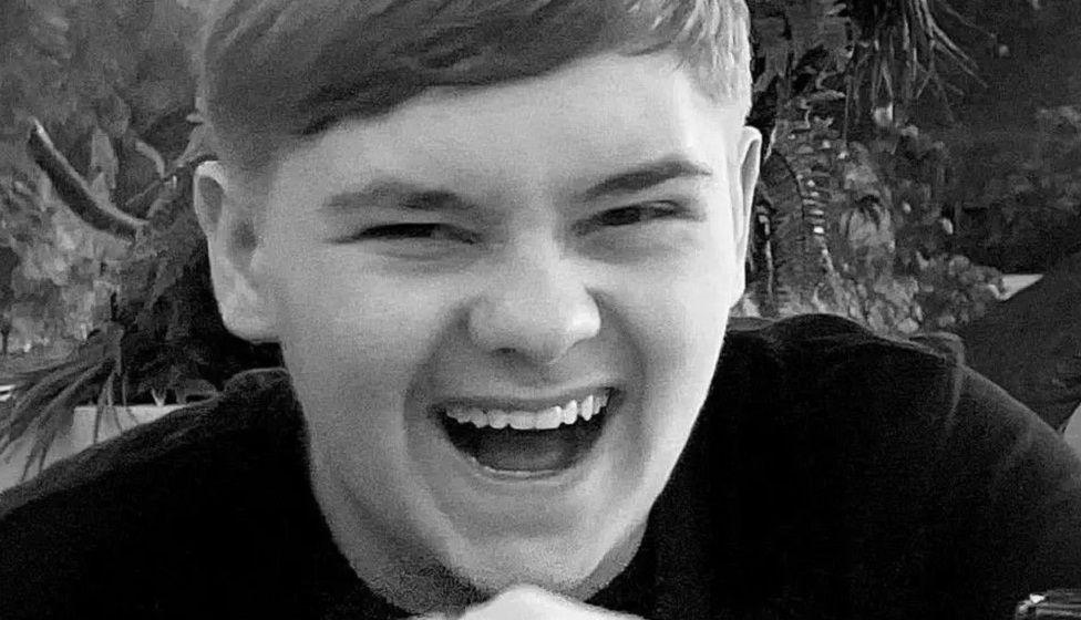 Harvey Willgoose laughing at the camera with his mouth open, wearing a black top. The photo is in black and white. 