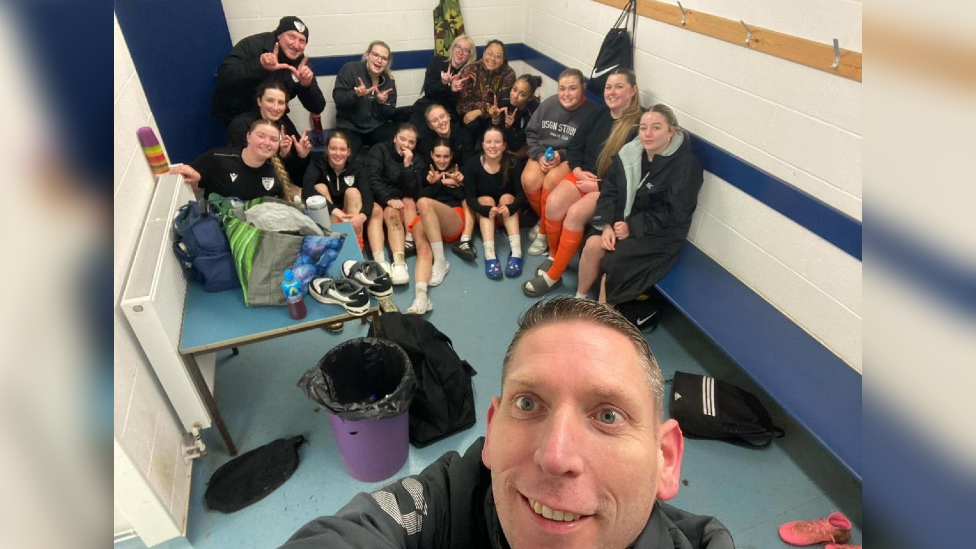 A selfie style photo with the club assistant coach at the bottom smiling at the camera and the girls behind. The team are in a locker room