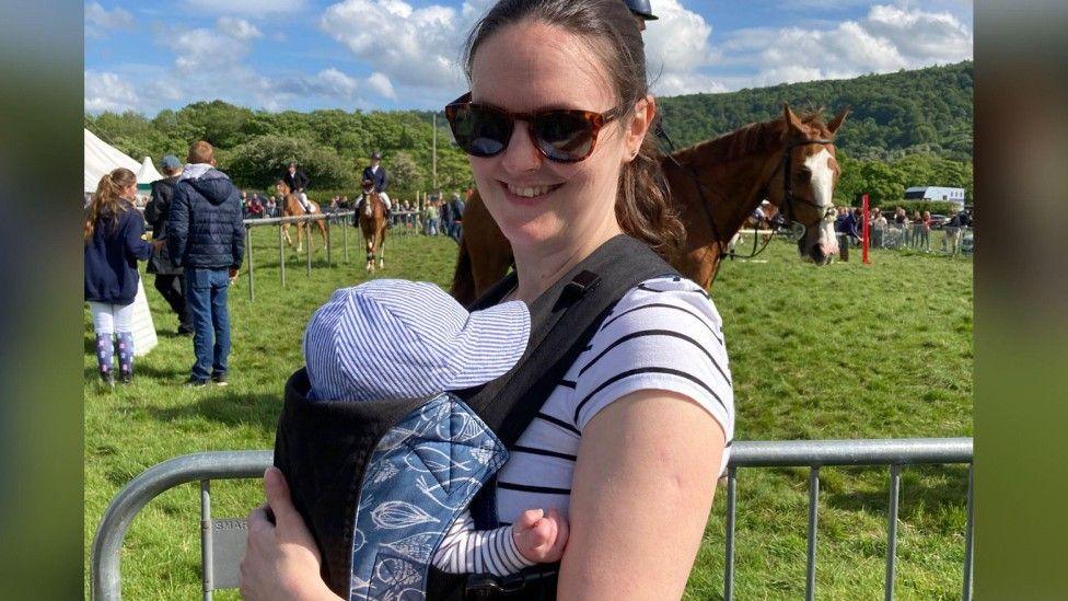 Gina, who has brown long hair in a ponytail and its wearing tortoiseshell sunglasses and a white and black striped t-shirt, pictured holding her baby son in a baby carrier