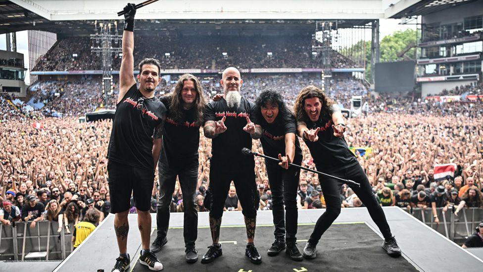 Five members of Anthrax pose in a row on stage in front of a huge stadium crowd
