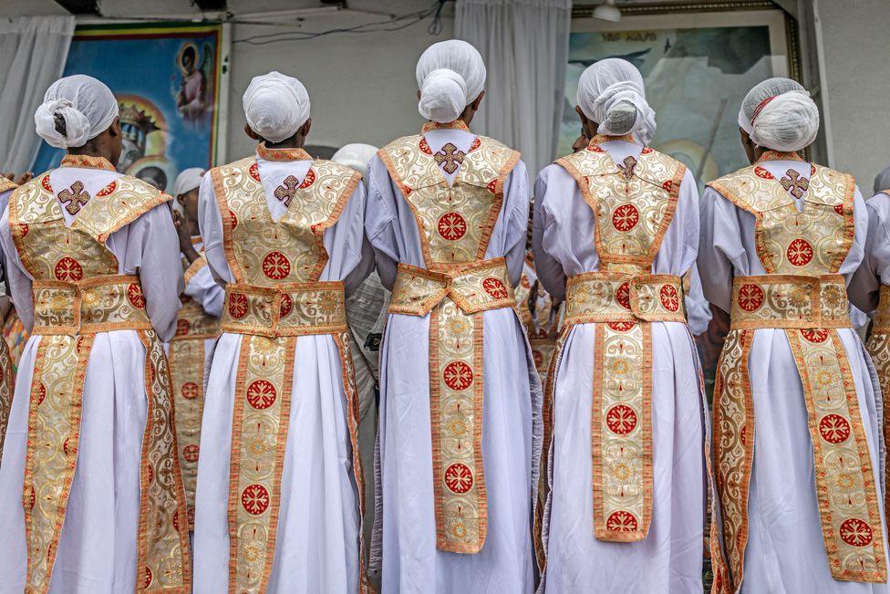 Five Orthodox deacons stand in a row. Seen from behind they are wearing white cassock robes with gold and red stoles. They are singing religious songs during a New Year church celebration at Entoto St Raguel Church in Addis Ababa, Ethiopia - Thursday 11 September 2025.