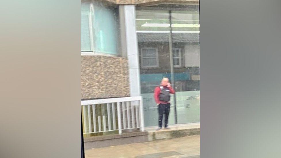 Blurry picture of a man wearing a tactical vest outside a business premises in Norwich. The man is seen talking on a mobile phone. 