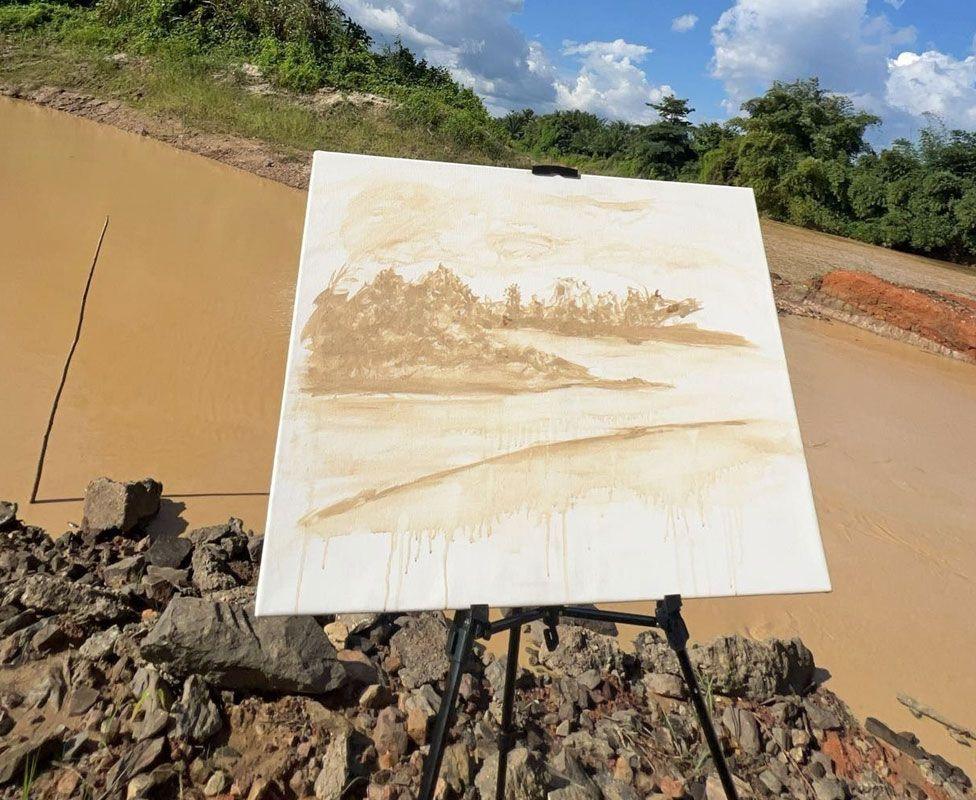 A painting of the Pra River using a yellow-brown colour on a white canvas, on a stand in front of the polluted river