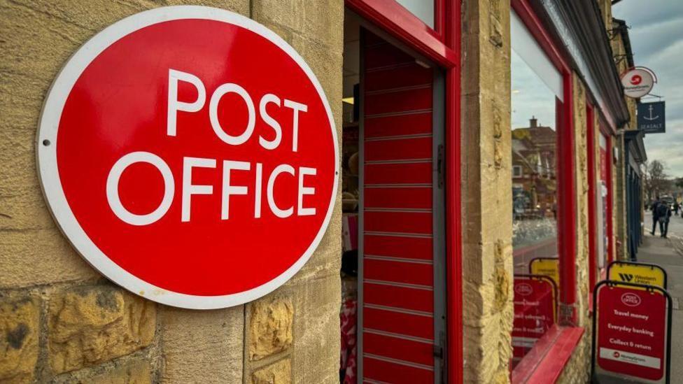 Exterior of a Post Office - red post office sign on a yellow stone wall, with shop windows and open door visible