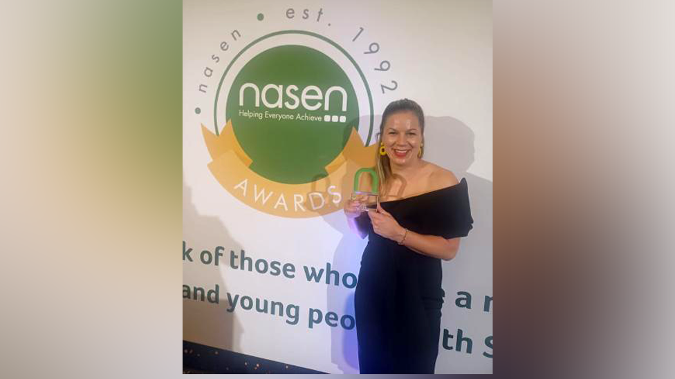 Michelle Madziak - a woman with long hair and wearing a black dress - holds up her award in front of a wall displaying the Nasen Awards logo.