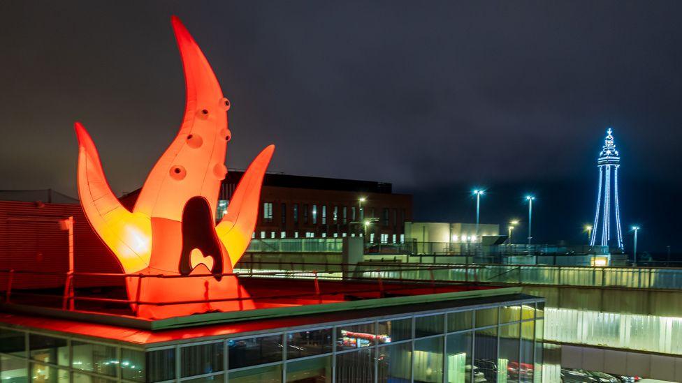 An orange inflatable monster on a rooftop in Blackpool on a dark night. It looks like a flame and has six bulbous eyes and a big black screaming mouth. Blackpool Tower can be seen in the distance to the right, lit up in blue