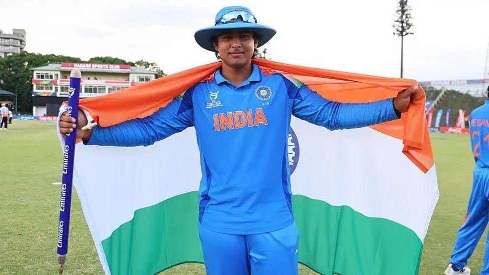 Vaibhav Sooryavanshi is standing facing the camera holding an Indian flag across his back in a Blue India strip. He is at a cricket ground and his holding a stump in his right hand