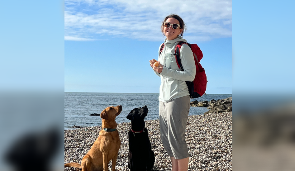 Dr Thirlwell standing on a pebble beach in front of the sea, wearing a light cream coloured zip hoody and a red backpack. She has light green cropped trousers, her brown hair tied back, and sunglasses as she smiles at the camera.
Looking up at her are two Labradors, one tan and one black, as they sit on the pebbles.