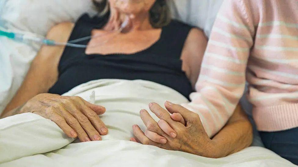 Two people hold hands. Their faces are out of the shot, but it is clear one person is in a hospital bed while the other stands be their side. The condition of their skin suggests the person in bed is much older than the person standing by their side. A tube can be seen attached to the older person suggesting they are sick.