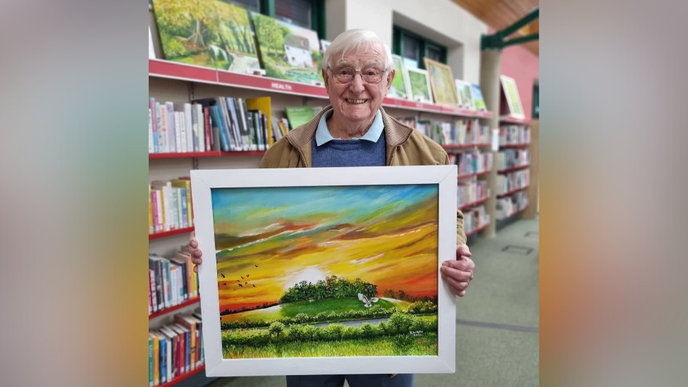 Robert Rose with library shelves behind him, holding a frames painting of a countryside sunset