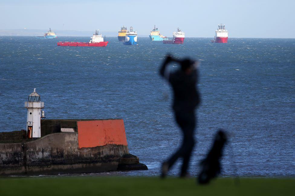 Silhouette of a golfer, with a lighthouse in the background, and offshore industry vessels in the sea.