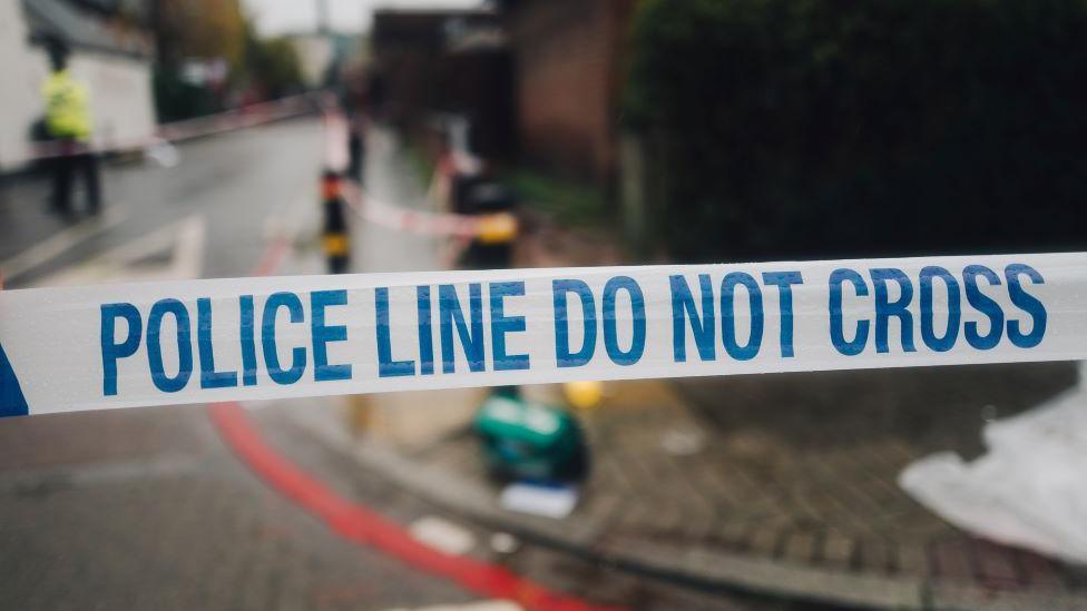 Stock image of police tape in front of what looks like a crime scene with emergency crews and officers 