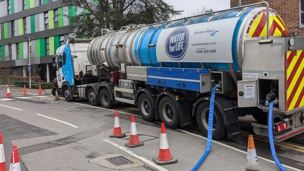 Water tanker outside green and green building with blue hoses coming from it onto the road which has red an white cones along it.