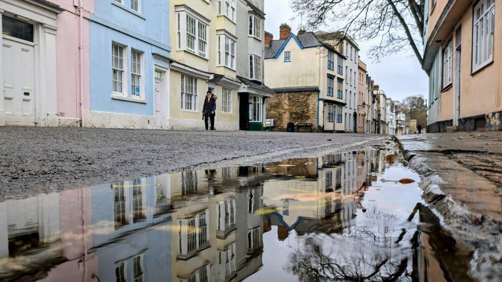 Flooding on a road with a cobbled pavement and pastel homes either side.