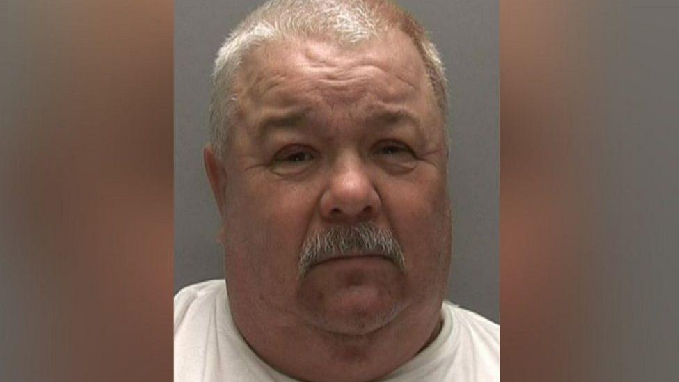 Police mugshot image of Gary Ward. He has a grey moustache and hair and is wearing a white top. He is pictured in front of a grey background and is squinting at the camera.