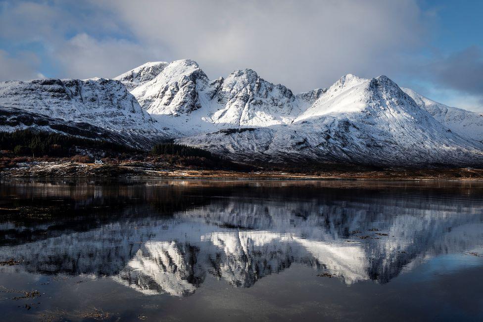 Mountain range peaks covered in snow, and shown in reflection in loch water.