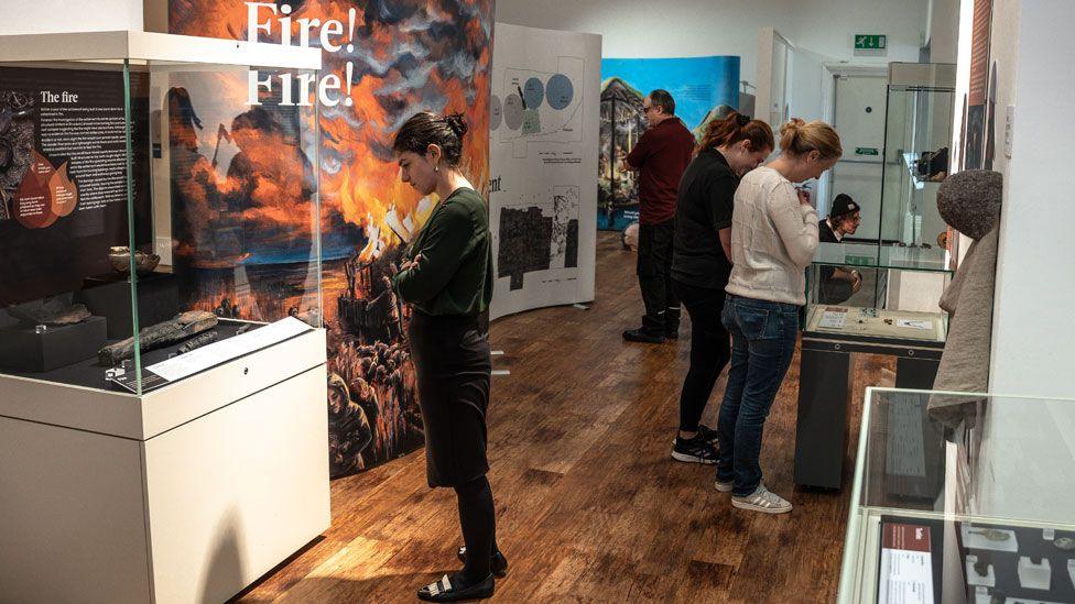 A woman in a dark top, skirt and tights, looking at the contents of an exhibition case, in front of a large display showing an artist's impression of a Bronze Age settlement going up in flames, with two men and two women looking at other displays at Peterborough Museum