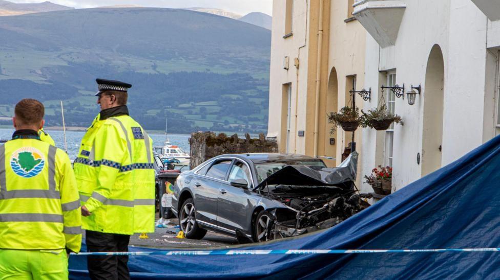 Two men in high vis, one wearing a police hat and jacket, stood next to a line of police tape. Beside them is a black car which is badly damaged and has crashed into a row of white houses. Behind is water, with a boat on it, and mountains.