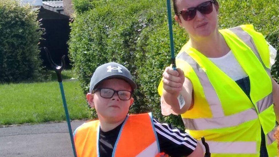 A boy aged about 9 wearing a black baseball cap, black framed glasses, a hi-vis orange waistcoat over a black T-shirt. To his left is a woman with her hair pulled back, wearing sunglasses, a yellow hi-vis jacket over a white T-shirt. Both have raised up blue-handled litter pickers in their right hands. Behind them is a hedge, grass and tarmacked road.
