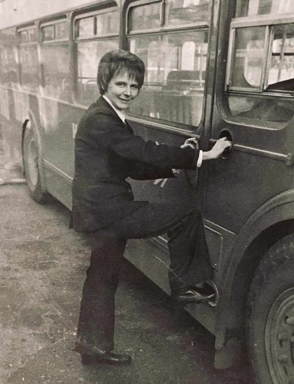 A black and white photo of Mary Curry standing next to a bus in 1973, one foot resting on the door. She is wearing a company uniform which includes a suit jacket and flared trousers. She has short hair.