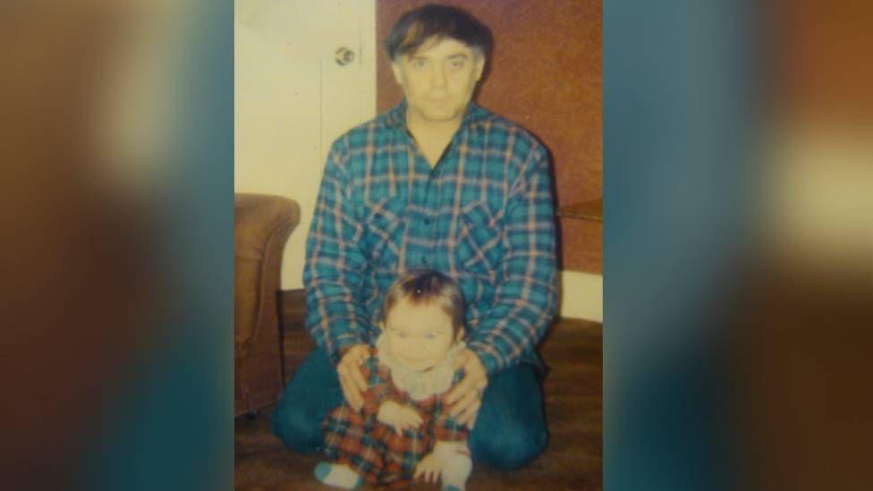 A old photo of a young baby wearing a tartan dress. She is being held by a man, her father, who is crouching down for the photo. He has black hair and has a blue chequered shirt on.