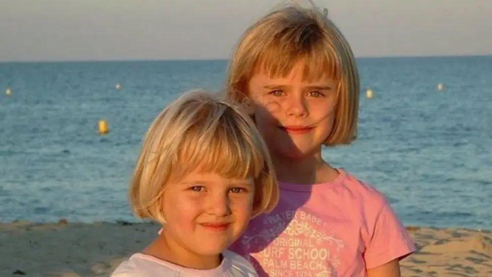 Laura and Gracie Nuttall standing in the sunshine on a beach as children. Both have blonde bobbed hair and are wearing pink tshirts