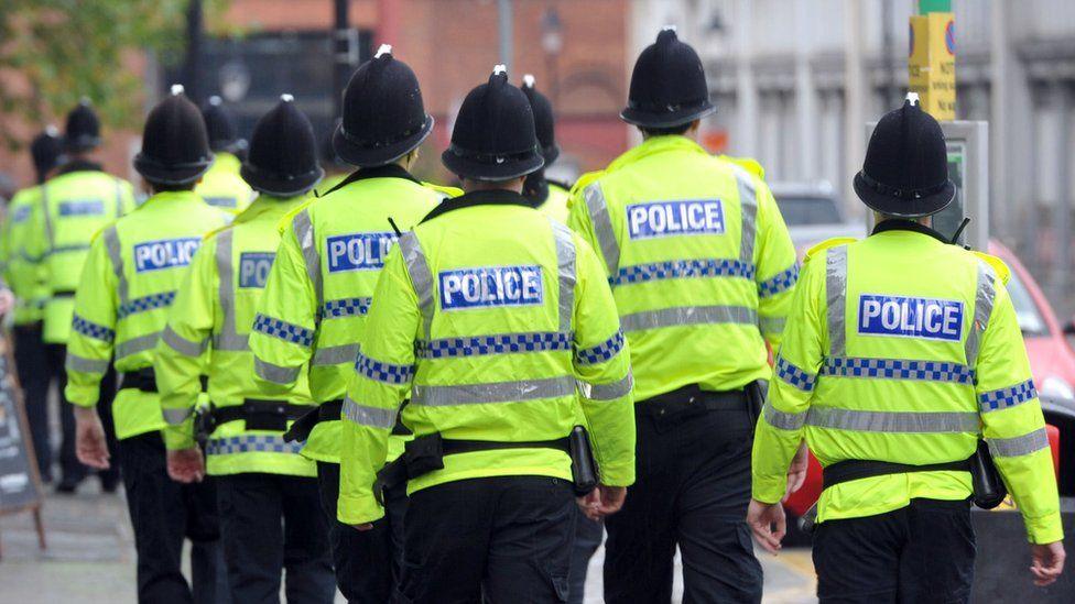 A group of Humberside Police officers marching down a street in Hull. They have their backs to the camera.  They are wearing fluorescent jackets with the word "police" on the back. They are all wearing traditional black and silver police helmets